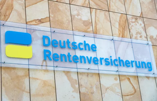 An einer hellen Steinwand hängt ein Glasschild mit der blauen Aufschrift "Deutsche Rentenversicherung". Links neben der Schrift ist ein Zeichen in Form von zwei Sprechblasen in gelb und blau. 