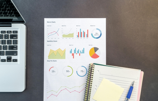 Arbeitsfläche mit einem geöffneten Laptop, einem Blatt mit verschiedenen bunten Diagrammen und einem Notizbuch