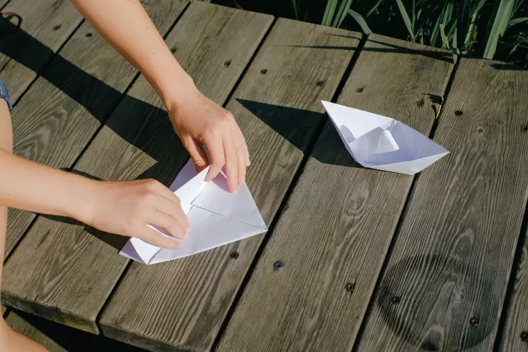 Zwei Hände falten ein Papierboot. Ein zweites Papierboot liegt fertig daneben.