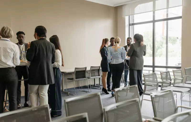 Zwei Gruppen von Personen stehen jeweils um einen Stehtisch herum und sprechen miteinander, im Vordergrund sind mehrere Stuhlreihen zu sehen