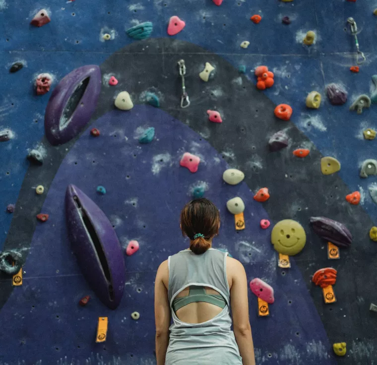 Eine Frau steht vor einer blauen Kletterwand mit bunten Klettergriffen und schaut nach oben. 