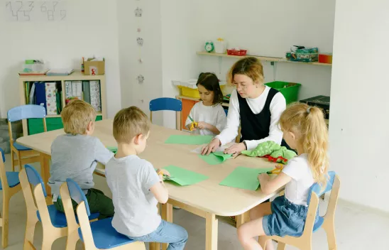 Eine Person sitzt mit mehreren Kindern an einem Tisch und bastelt etwas Eine Person sitzt mit mehreren Kindern an einem Tisch und bastelt etwas