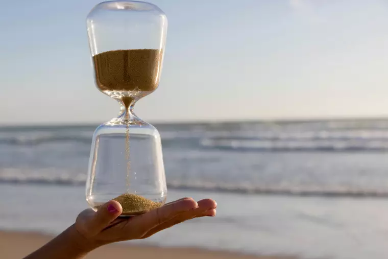 Eine Hand hält eine Sanduhr. Im Hintergrund sieht man Strand und Meer.