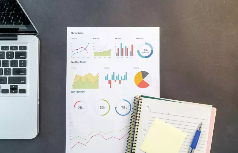 Arbeitsfläche mit einem geöffneten Laptop, einem Blatt mit verschiedenen bunten Diagrammen und einem Notizbuch