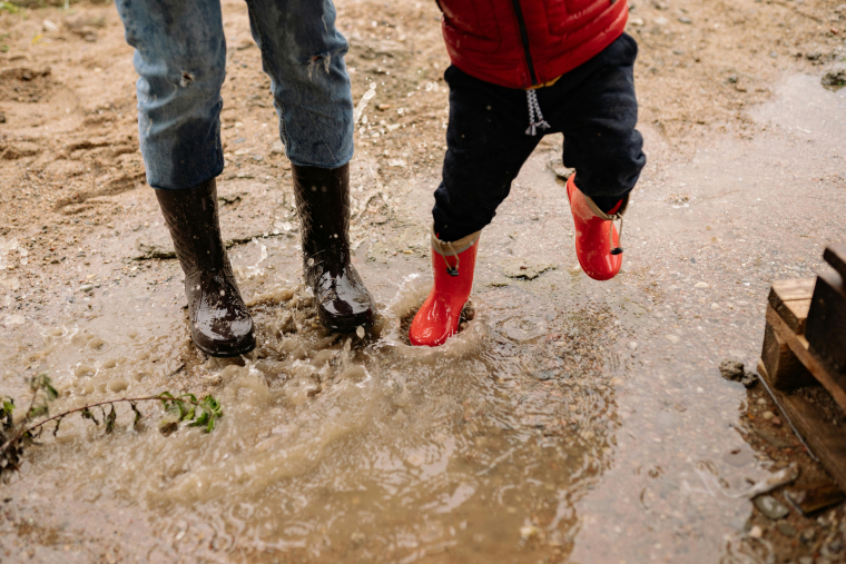 Beine zweier Personen, eine erwachsen, eine im Kindesalter, mit Gummistiefeln bekleidet in einer Pfütze springend.
