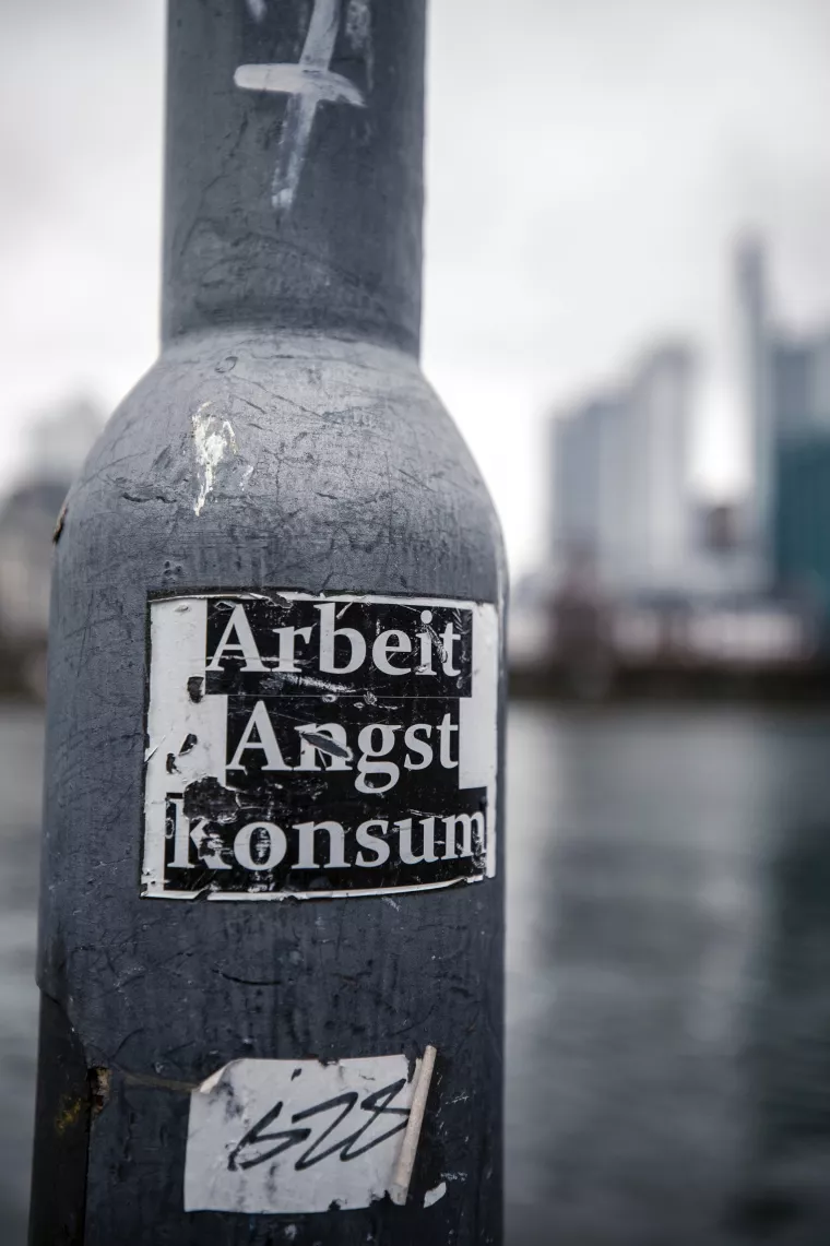 Ein zerkratzter Sticker mit der Aufschrift "Arbeit, Angst, Konsum" klebt an einer Laterne. Das Foto ist Schwarzweiß. 