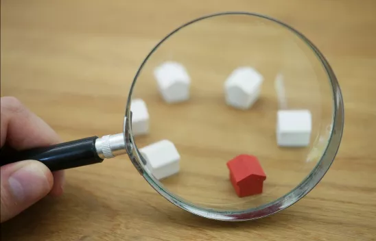 Eine Lupe wird von einer Hand gehalten und zu sehen sind fünf kleine weiße Holzhäuser sowie ein rotes kleines Holzhaus Eine Lupe wird von einer Hand gehalten und zu sehen sind fünf kleine weiße Holzhäuser sowie ein rotes kleines Holzhaus