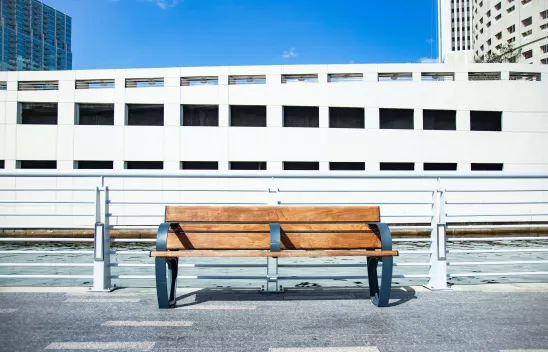 Foto einer Holzbank am Straßenrand. In der Mitte der Bank ist eine dritte Lehne, die das Liegen auf der Bank unmöglich macht und ein Beispiel für wohnungslosenfeindliche Architektur ist. 