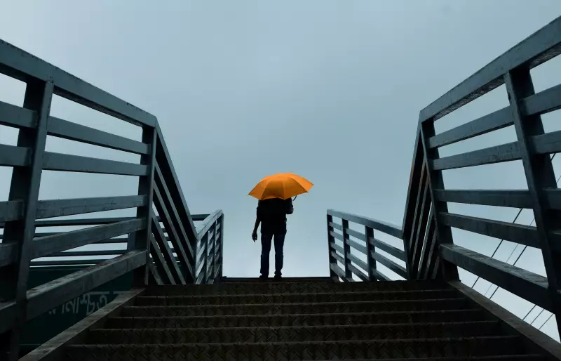 Eine Person steht auf der obersten Stufe einer Treppe und hält vor grauem Himmel einen orangefarbenen Regenschirm; sie ist von hinten als Silhouette erkennbar.