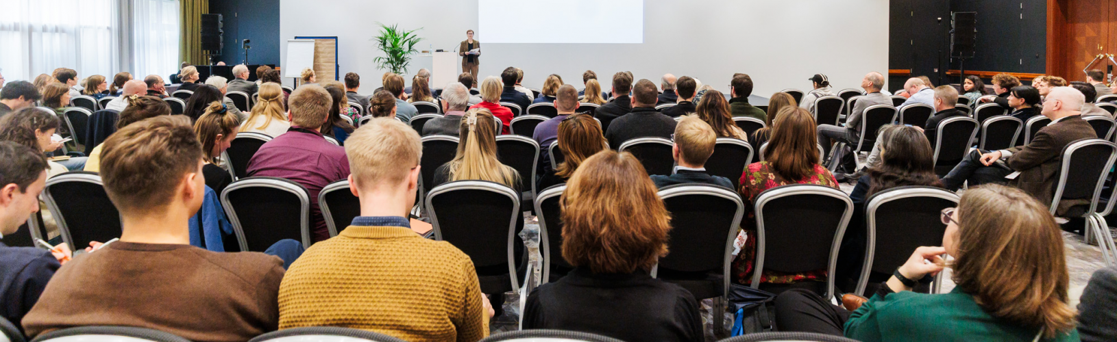 Blick in einen Konferenzsaal. Auf der Bühne steht eine Person, die einen Vortrag hält.