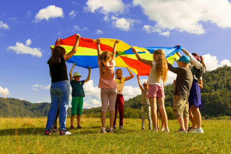 Gruppe von Kindern und Erwachsenen, die auf einer Wiese mit einer Regenbogenplane spielen