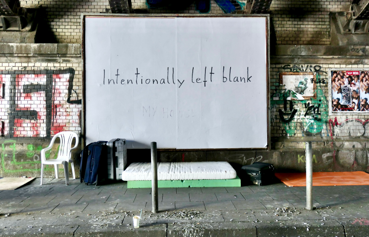 Auf dem Bürgersteig unter einer Brücke liegen Matratzen, Koffer und links steht ein Stuhl. Dahinter auf einer großen weißen Plakatwand steht die Aufschrift "Intentionally left blank".