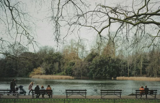 In einem Park sitzen Menschen unterschiedlichen Alters auf Bänken. Menschen mit Kinderwagen gehen vorbei. Alle sind aus der Ferne von hinten zu sehen. 