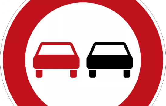 Ein Überholverbotsschild: Ein roter Kreis in dem links ein rotes Auto und rechts ein schwarzes Auto abgebildet sind. 