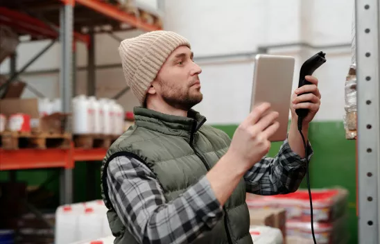 Person mit Mütze und Weste scannt Ware in einem Lager mit Tablet und Handscanner Person mit Mütze und Weste scannt Ware in einem Lager mit Tablet und Handscanner
