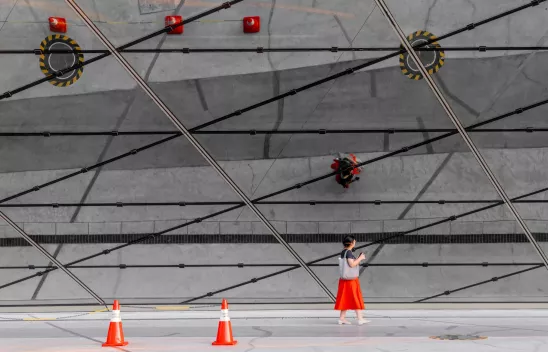 Eine Person mit rotem Rock läuft vor einer Glasfassade, auf der sich die Struktur des Straßenbelags und rote Baustellenkegel spiegeln. Eine Person mit rotem Rock läuft vor einer Glasfassade, auf der sich die Struktur des Straßenbelags und rote Baustellenkegel spiegeln.