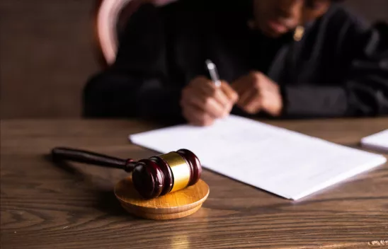 Im Vordergrund liegt ein Gerichtshammer auf einer Holzscheibe auf einem Tisch. Im Hintergrund sieht man, wie eine Person, die am Tisch sitzt, etwas auf ein Blatt Papier schreibt. Im Vordergrund liegt ein Gerichtshammer auf einer Holzscheibe auf einem Tisch. Im Hintergrund sieht man, wie eine Person, die am Tisch sitzt, etwas auf ein Blatt Papier schreibt.