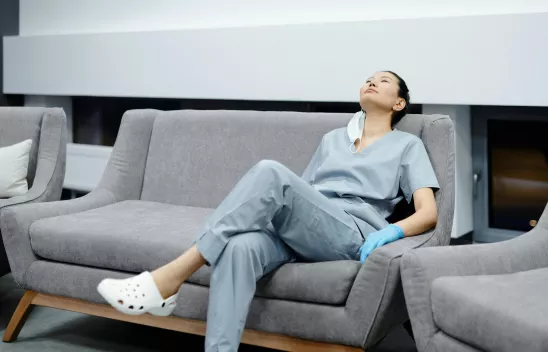 Eine Person in Krankenpfleger-Kleidung sitzt auf einem Sofa und sieht erschöpft aus Eine Person in Krankenpfleger-Kleidung sitzt auf einem Sofa und sieht erschöpft aus