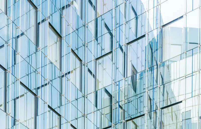 Die Glasfassade eines Gebäudes spiegelt eine angrenzende Fassade, wodurch ein  geometrisches Muster aus Linien und Reflexionen entsteht.