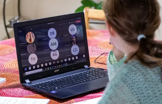 Eine Person sitzt an einem Laptop, auf dem Bildschirm werden Kacheln einer Videokonferenz angezeigt Eine Person sitzt an einem Laptop, auf dem Bildschirm werden Kacheln einer Videokonferenz angezeigt