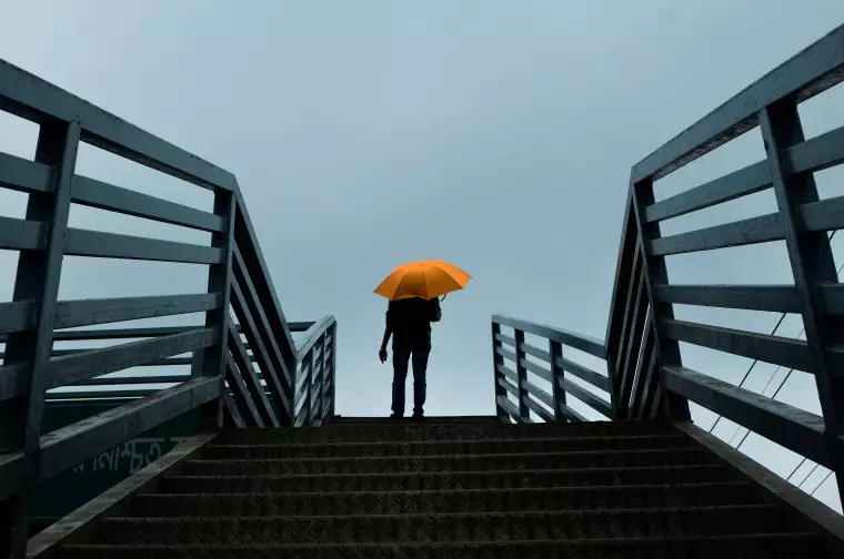 Eine Person steht auf der obersten Stufe einer Treppe und hält vor grauem Himmel einen orangefarbenen Regenschirm; sie ist von hinten als Silhouette erkennbar.