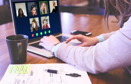 Eine Person sitzt an einem Laptop, auf dem Bildschirm werden Kacheln einer Videokonferenz angezeigt. Neben dem Laptop sind eine Tasse und ein Blatt Papier mit Stift zu sehen. Eine Person sitzt an einem Laptop, auf dem Bildschirm werden Kacheln einer Videokonferenz angezeigt. Neben dem Laptop sind eine Tasse und ein Blatt Papier mit Stift zu sehen.