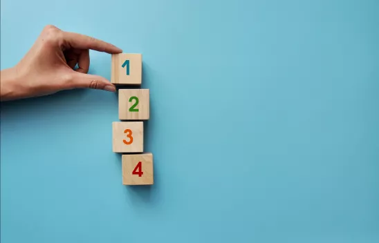 Auf hellblauem Hintergrund liegen vier Würfeln mit den Zahlen von eins bis vier farblich beschriftet. Eine hand hält mit zwei Fingern den ersten Würfel mit der Eins fest. 