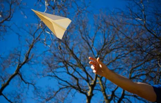 Ein ausgestreckter Arm lässt einen Papierflieger vor blauem Himmel und blätterlosen Bäumen fliegen. Ein ausgestreckter Arm lässt einen Papierflieger vor blauem Himmel und blätterlosen Bäumen fliegen.