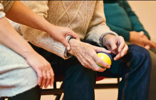 Blick auf zwei Personen, die nebeneinander sitzen. Eine jüngere Person hält die Hand einer älteren Person, in ihrer Hand befindet sich ein kleiner Ball. Blick auf zwei Personen, die nebeneinander sitzen. Eine jüngere Person hält die Hand einer älteren Person, in ihrer Hand befindet sich ein kleiner Ball.