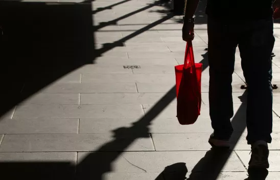 Person mit roter Einkaufstasche, von hinten fotografiert, Schatten von Laternen und der Person auf Betonboden sichtbar Person mit roter Einkaufstasche, von hinten fotografiert, Schatten von Laternen und der Person auf Betonboden sichtbar