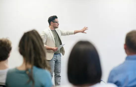 Eine Person steht vorne und präsentiert etwas, im Vordergrund sind verschiedene andere Menschen zu sehen, die dem Vortrag folgen. Eine Person steht vorne und präsentiert etwas, im Vordergrund sind verschiedene andere Menschen zu sehen, die dem Vortrag folgen.