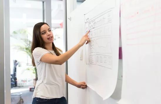 Eine Person steht vor einer Tafel und zeigt etwas auf einem Blatt Papier