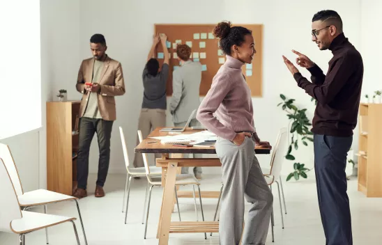 In einem Raum sind mehrere Personen zu sehen, zwei Personen beschäftigen sich mit einer Pinnwand, im Vordergrund unterhalten sich zwei Personen In einem Raum sind mehrere Personen zu sehen, zwei Personen beschäftigen sich mit einer Pinnwand, im Vordergrund unterhalten sich zwei Personen