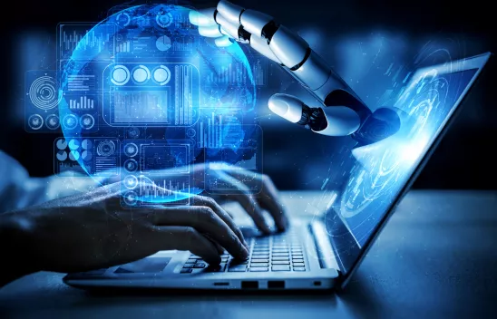 Aus einem Laptopbildschirm kommt eine Roboterhand, die eine digitale blaue Kugel berührt. Auf die Tastatur des Laptops tippen zwei Hände.