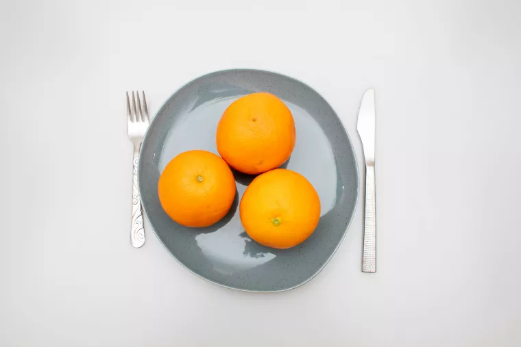 Drei Orangen auf einem Teller, rechts und links davon liegen Messer und Gabel.