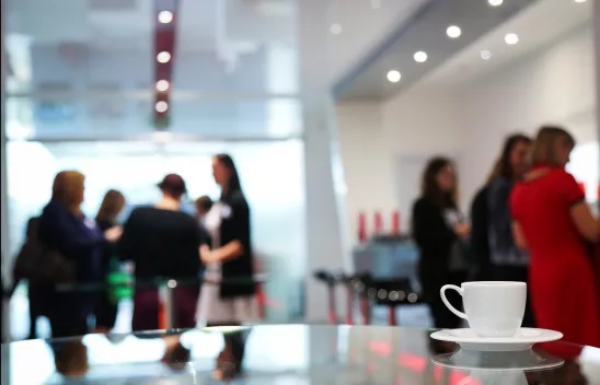 Im Vordergrund ein Stehtisch, auf dem eine kleine Kaffeetasse zu sehen ist, im Hintergrund sind Personen in Gruppen zu sehen, die miteinander sprechen Im Vordergrund ein Stehtisch, auf dem eine kleine Kaffeetasse zu sehen ist, im Hintergrund sind Personen in Gruppen zu sehen, die miteinander sprechen
