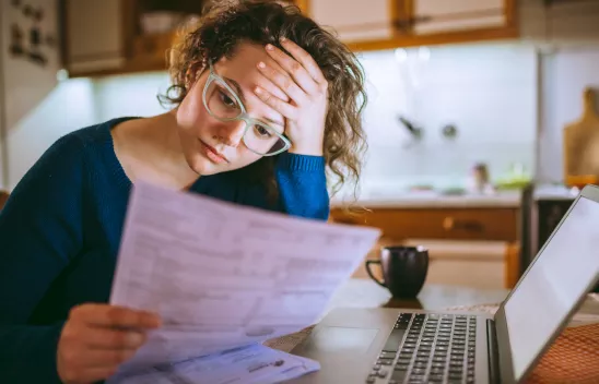 Eine Frau hat ihren Kopf auf einer Hand aufgestützt und sitzt vor einem Laptop. Sie hat ein Formular oder eine Rechnung in der anderen Hand und schaut darauf. 