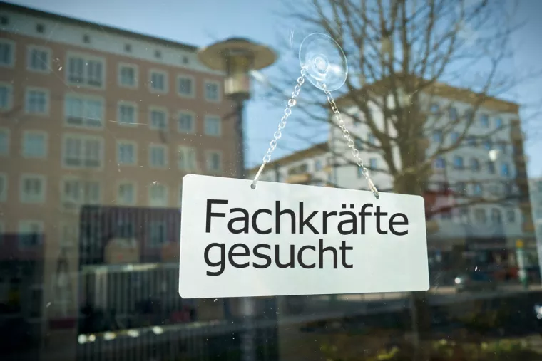 Auf einer Glasscheibe hängt ein Schild mit der Aufschrift "Fachkräfte gesucht". Es spiegeln sich Häuser im Hintergrund in der Scheibe.