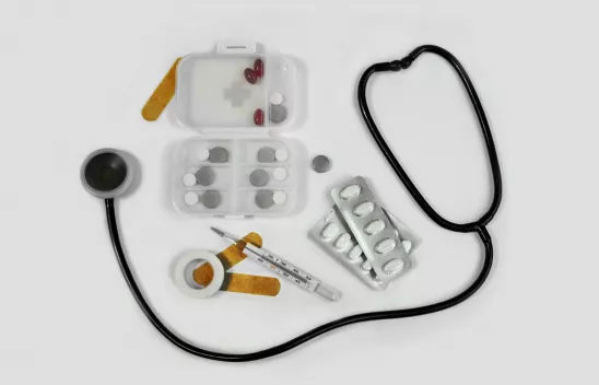 Fotografie von Pillendosen, Pflaster, Fieberthermometer und Stethoskop vor einem weißen Hintergrund.