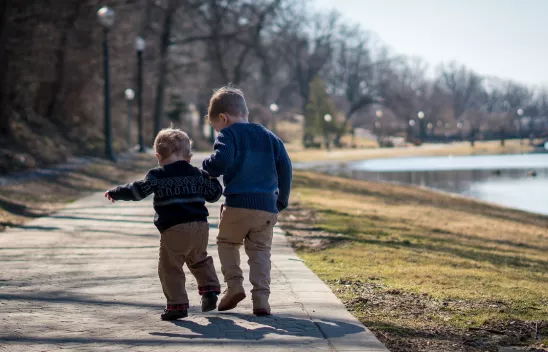 Zwei Kinder stehen auf einer Promenade, man sieht sie von hinten und sie halten sich an den Händen. Rechts davon ist eine Wiese und ein See zu sehen. Zwei Kinder stehen auf einer Promenade, man sieht sie von hinten und sie halten sich an den Händen. Rechts davon ist eine Wiese und ein See zu sehen.