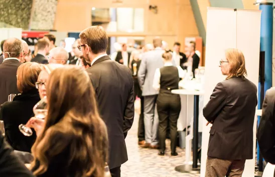 Verschiedene Gruppen stehen in einem Raum an Stehtischen und unterhalten sich Verschiedene Gruppen stehen in einem Raum an Stehtischen und unterhalten sich