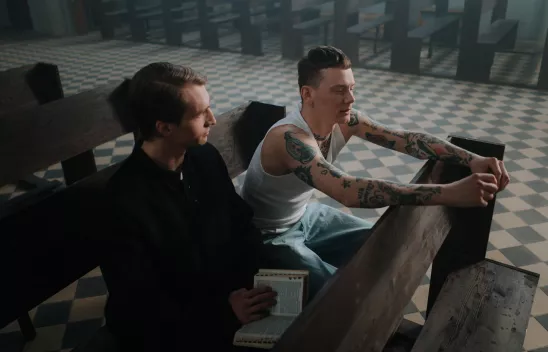 Ein Priester mit einer Bibel im Schoß sitzt neben einem Mann mit Tanktop und Tattoos auf einer Kirchenbank in einer leeren Kirche. Ein Priester mit einer Bibel im Schoß sitzt neben einem Mann mit Tanktop und Tattoos auf einer Kirchenbank in einer leeren Kirche.