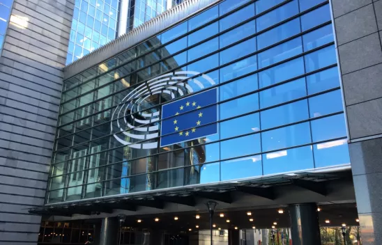 Ein Gebäude mit einer Glasfront, auf der mittig ein Konferenzsaal in weiß und die europäische Flagge (blauer Hintergrund mit einem gelben Sternenkreis) aufgeklebt sind. Ein Gebäude mit einer Glasfront, auf der mittig ein Konferenzsaal in weiß und die europäische Flagge (blauer Hintergrund mit einem gelben Sternenkreis) aufgeklebt sind.