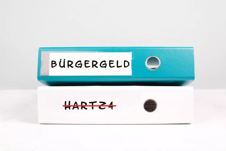 Ein blauer Ringordner mit der seitlichen Aufschrift "Bürgergeld" liegt auf einem weißen Ringordner mit der in rot durchgestrichenen Aufschrift "Hartz 4".