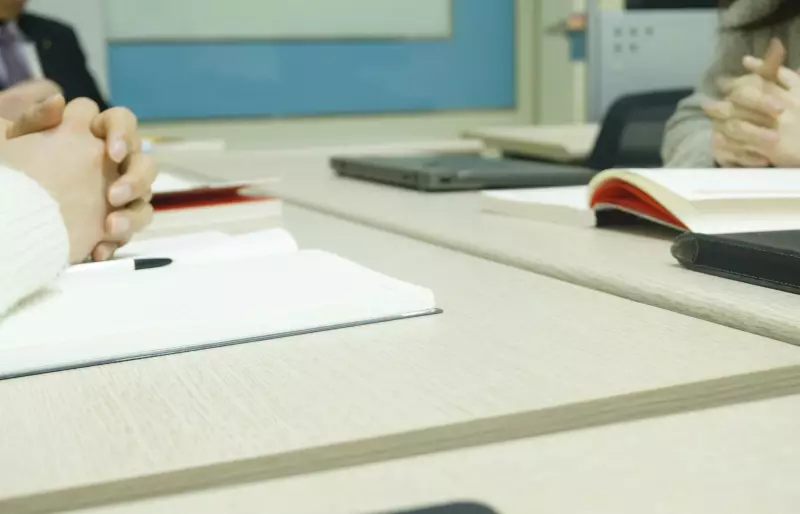 Personen sitzen an einem Tisch in einer Besprechung; vor ihnen liegen Notizbuch, Stift, Laptop und Unterlagen.