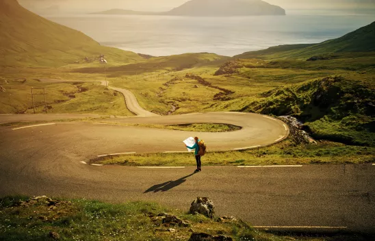 Eine Person mit einer Landkarte in den Händen steht auf einer Serpentinenstraße zwischen grünen Wiesen, die am Meer mündet. 