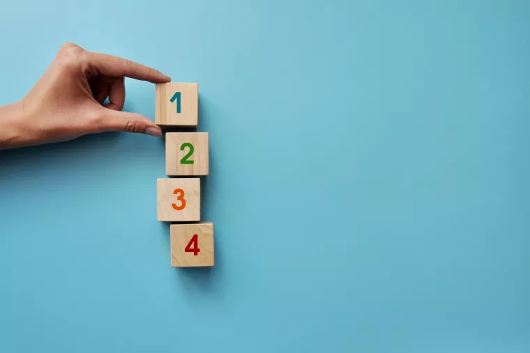 Auf hellblauem Hintergrund liegen vier Würfeln mit den Zahlen von eins bis vier farblich beschriftet. Eine hand hält mit zwei Fingern den ersten Würfel mit der Eins fest. 