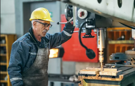 Ein älterer Arbeiter mit Schutzkleidung und gelbem Schutzhelm arbeitet an einer Metallschneidemaschine in einer Halle.