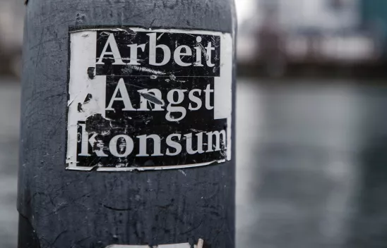 Ein zerkratzter Sticker mit der Aufschrift "Arbeit, Angst, Konsum" klebt an einer Laterne. Das Foto ist Schwarzweiß. Ein zerkratzter Sticker mit der Aufschrift "Arbeit, Angst, Konsum" klebt an einer Laterne. Das Foto ist Schwarzweiß.