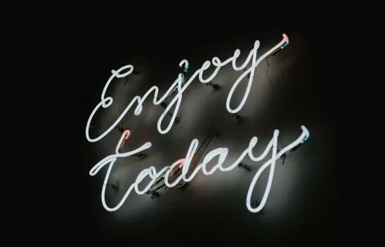 Ein Schriftzug in weißem Neonlicht mit dem Titel "Enjoy today" leuchtet in weiß vor einem dunklen Hintergrund.